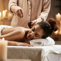 A woman face down on a towel on a massage table with a person dropping oil on her back, They are surrounded by candles.