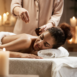 A woman face down on a towel on a massage table with a person dropping oil on her back, They are surrounded by candles.